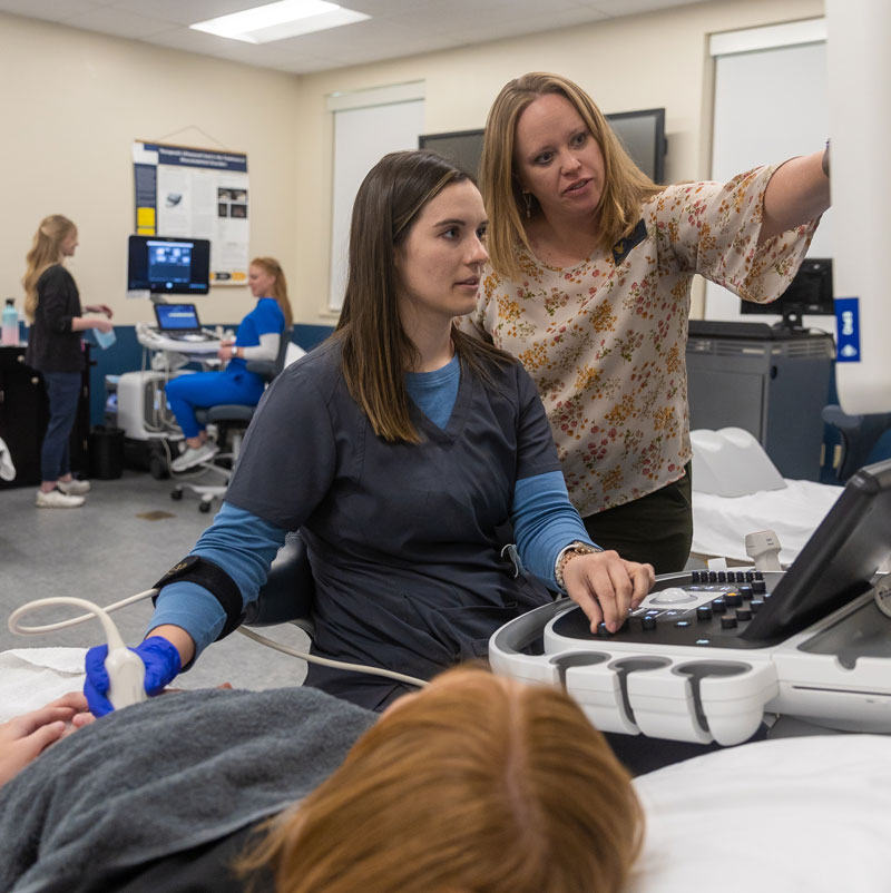 photo of a faculty member pointing at a screen while a student does an ultrasound on a partner in the Sonography Lab on campus.