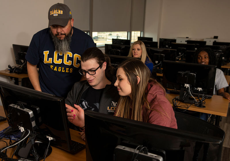 photo of students in a computer lab. Three students are looking at one screen. There are two more students in the background looking at computers.