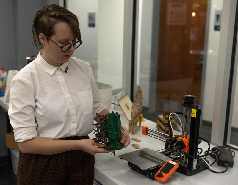 photo of female student near a 3D printer in the Library Markerspace. She is holding a 3D printed Groot figure.