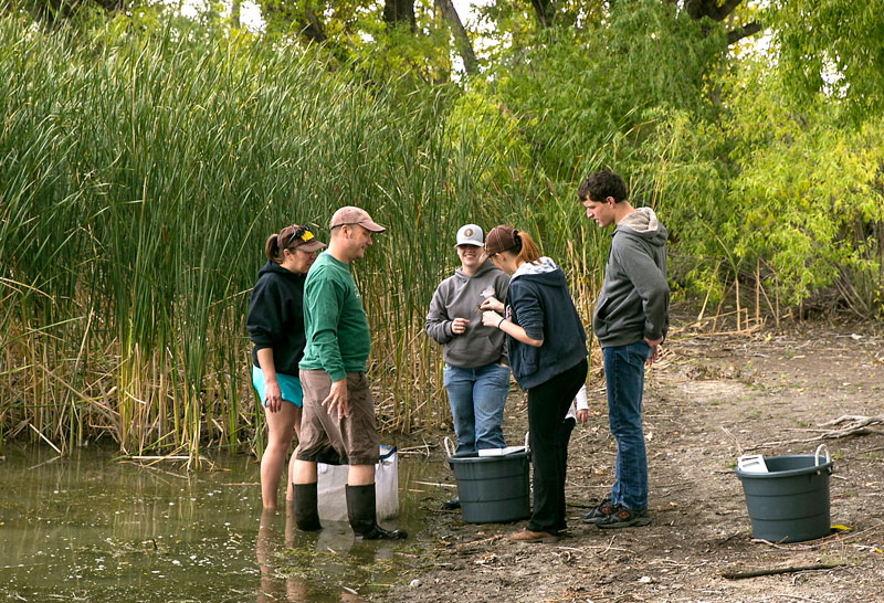 Dr. Zac Roehrs (in green) outdoors with students collecting samples from the pond on campus