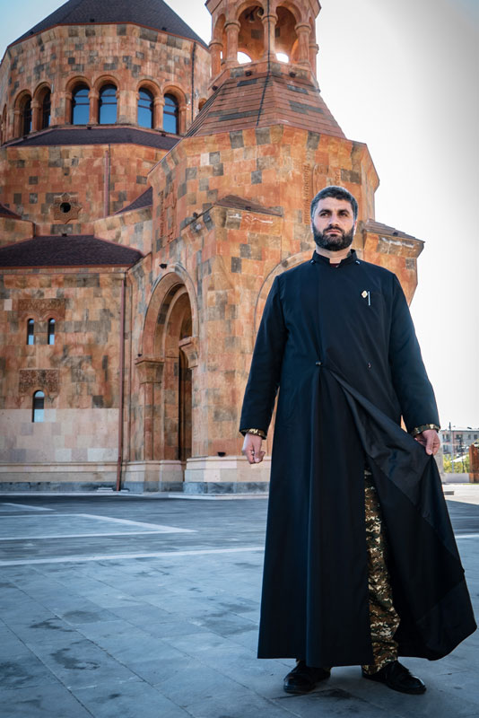 Photo by Ezras Tellalian with a man standing in front of a church dressed as a priest in a robe while holding it open to show his militray fatigues underneath.