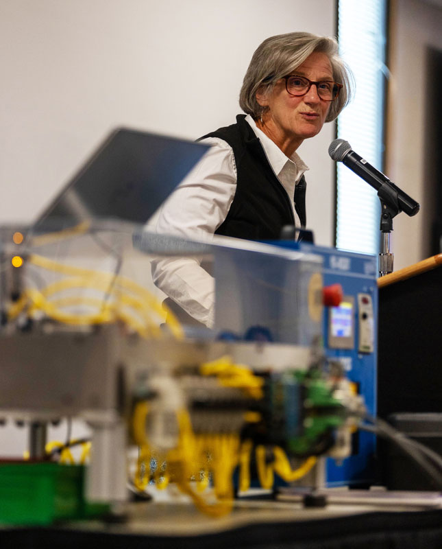 photo of Kari Brown-Herbst talking at a podium about manufacturing with a plexiglass box with wires a buttons in front of her.