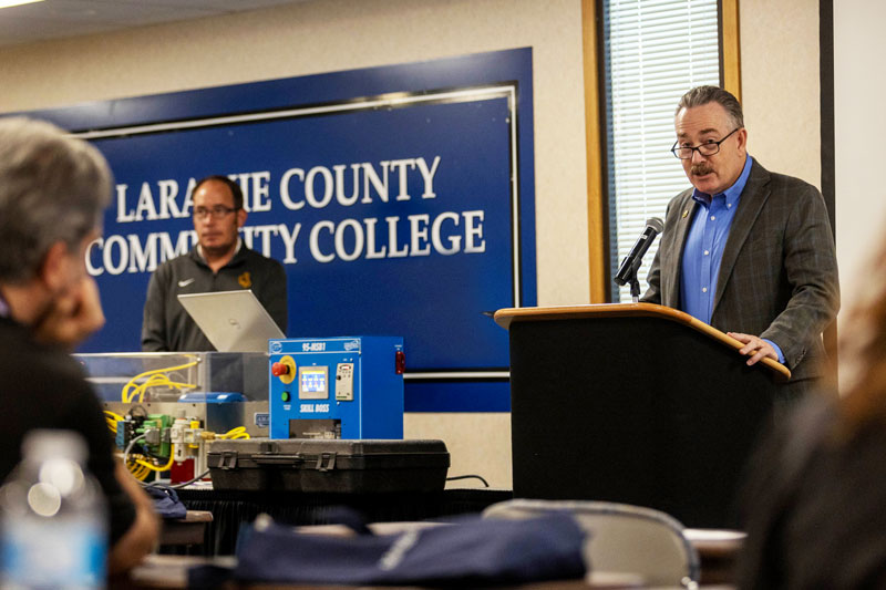 photo of Cheyenne Mayor Patrick Collins talking at a podium about manufacturing with a plexiglass box with wires a buttons in front of him. There are people in the audience listening to him.