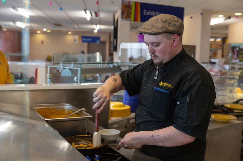 photo of a male cook in the LCCC Dining Hall mixing a sauce in a skillet. He's wearing a uniform and hat.
