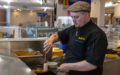 photo of cook in LCCC Dining Hall scraping a pan with a spatula as it cooks over a stove.