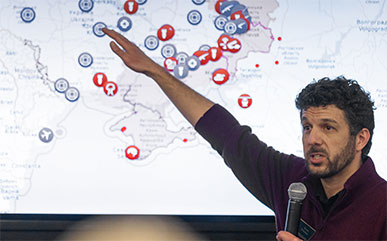 photo of Ezras Tellalian in front of a projected map of Ukraine holding a microphone. He's giving a talk about the war in Ukraine.