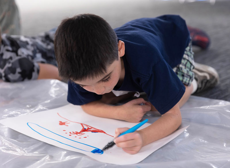 A student drawing with paintbrush markers on a big paper on the floor SEEK class