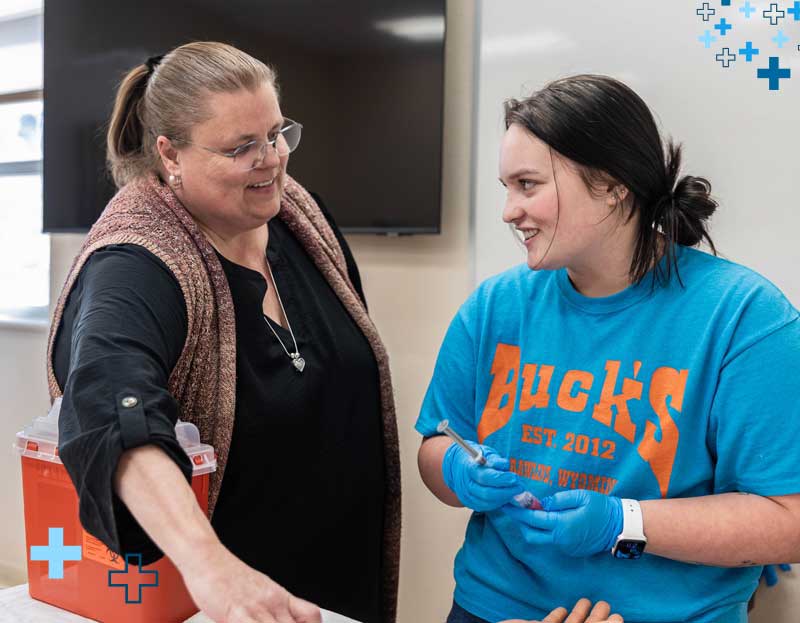 faculty member and student in a Health Sciences lab classroom with gloves on and a sharps container in front of them