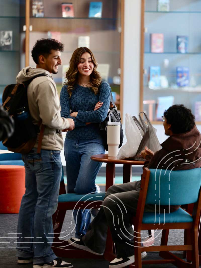 Two students standing and talking with a student sitting at a table in the library on campus