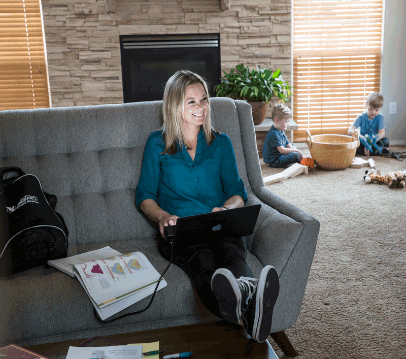 photo of student sitting on a couch at home with a laptop. Children are playing in the background.