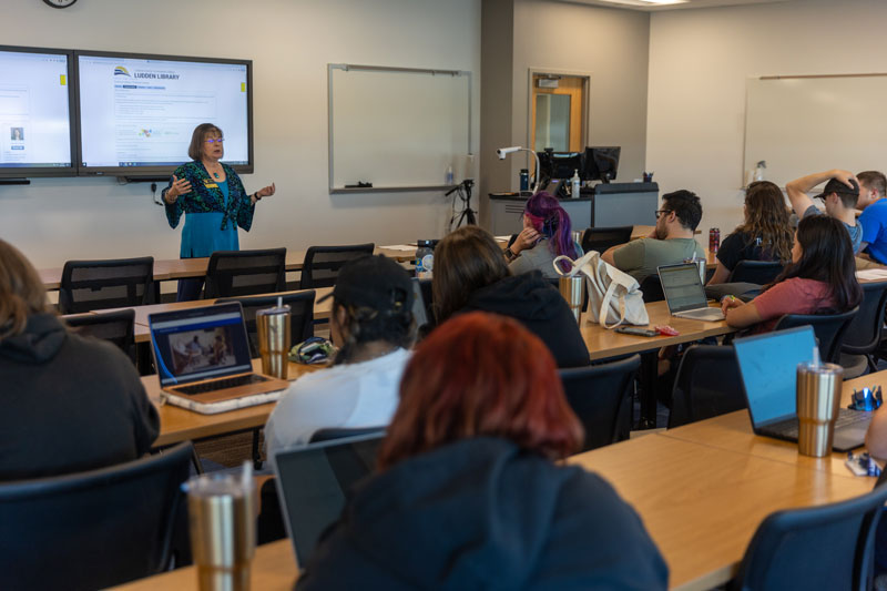 Photo of a class in a classroom in the Clay Pathfinder Building with two screens up front. Students sit a tables with laptops.