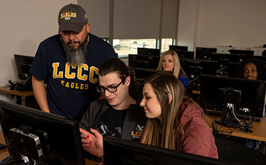 photo of students in a computer lab. Three students are looking at one screen. There are two more students in the background looking at computers.