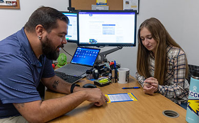 photo of LCCC advisor talking to female student in office. They are looking at information on a printed page with computer screens in the background.