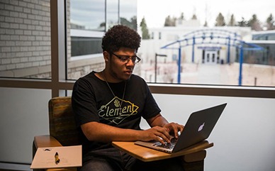 Photo of student sitting in a chair with a laptop and paper and pen by window in the Clay Pathfinder Building.