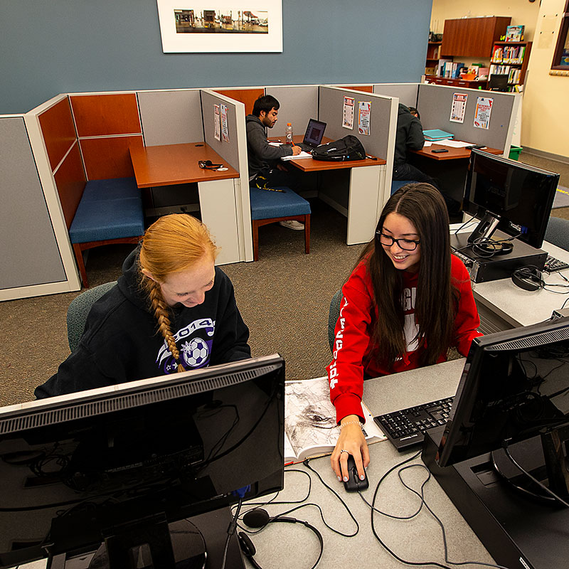 photo of two female students working at computers with a male student working at a desk behind them