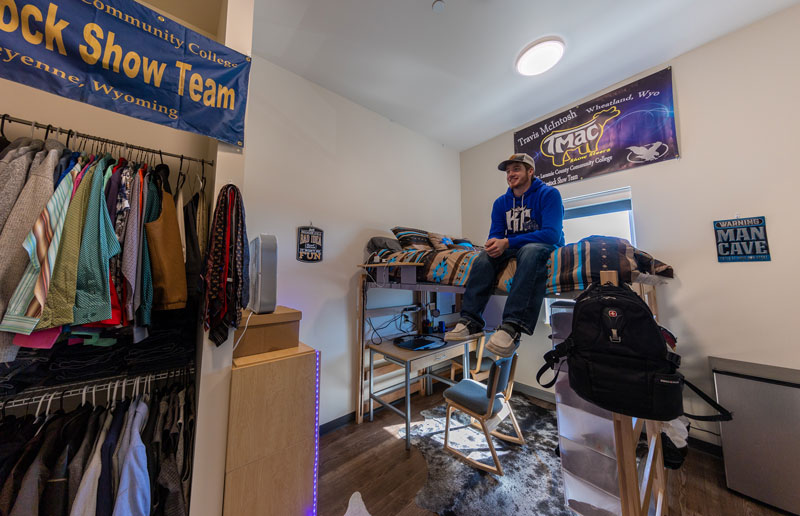 Male student sitting on a lofted bed in a dorm room in Gold Hall. The bed has a desk and computer under it, and you can see part of the closet in the room.