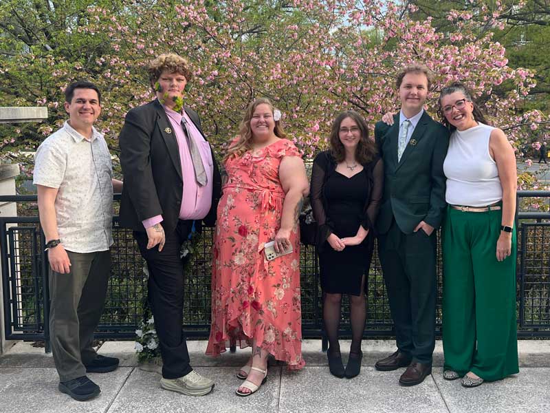 A group photo of five speech and debate students with their advisor dressed up for a competition. They are standing outside with trees in the background.