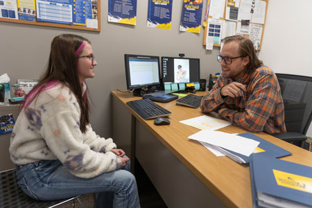 Photo of a student sitting across a desk from Student Success Coach Tyler Garrison. They are in his office with papers on a bulletin board behind them and laying on the desk near a computer.