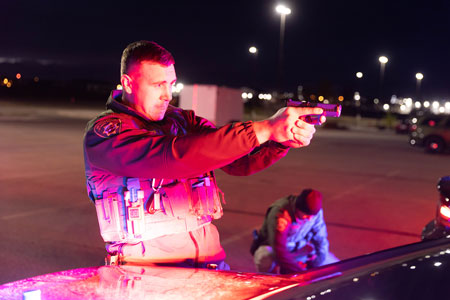 photo of law enforcement training simulation where an officer in training is aiming a firearm in the distance and highlighted by the light from police car.