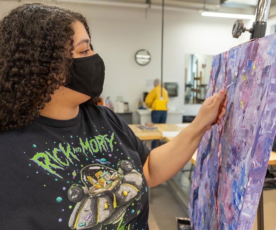 photo of a female student wearing a face mask painting on an easel in a classroom.