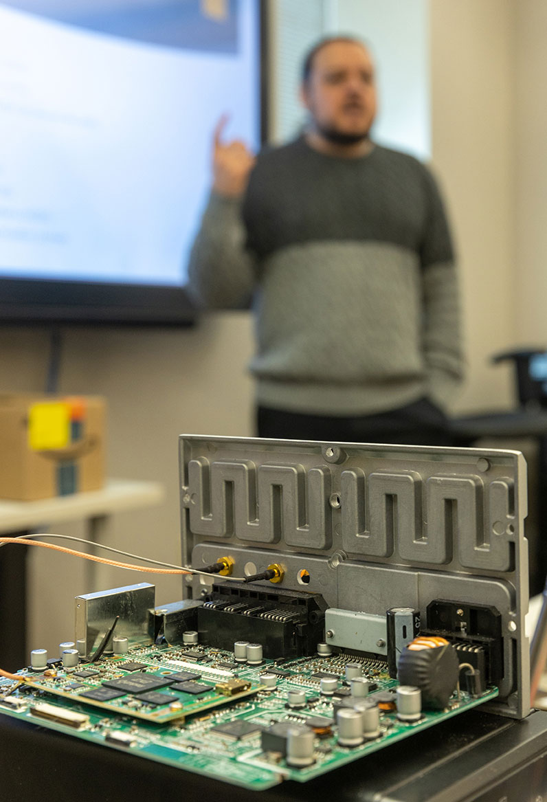 Photo of the inside of a computer in the foeground with a faculty member speaking next to a screen in the background.