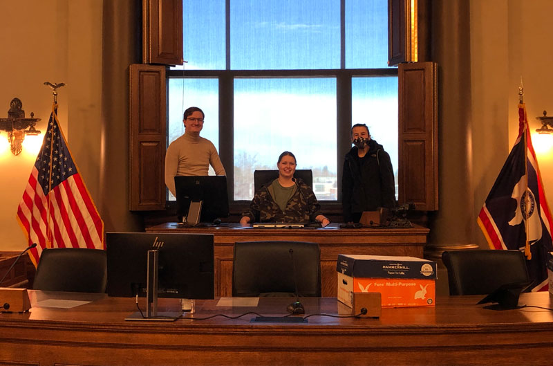photo of legislative intern sitting at the main desk in the legislative office surrounded by two other people.