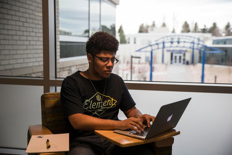 photo of student in a chair working on a laptop with a pad of paper and a pen..