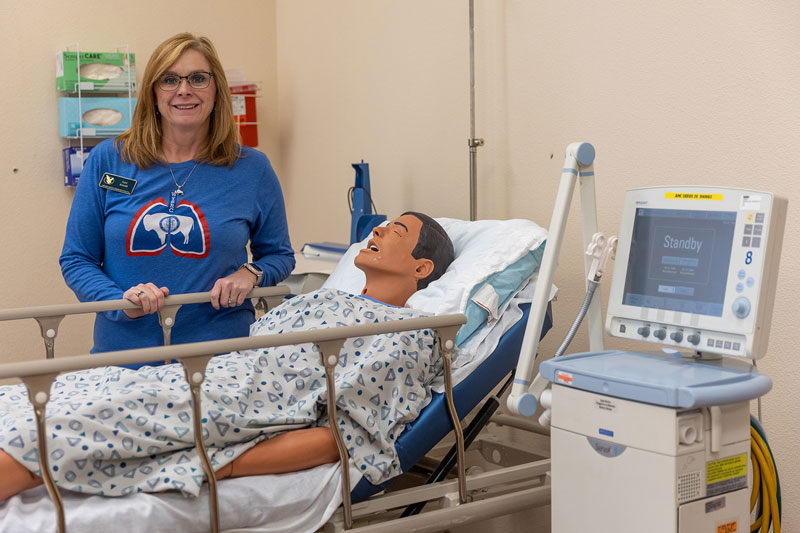 Photo of Lori Arnold standing behind a hospital bed in the LCCC Health Sciences nursing simulation lab. The bed has a simulation patient laying in it.