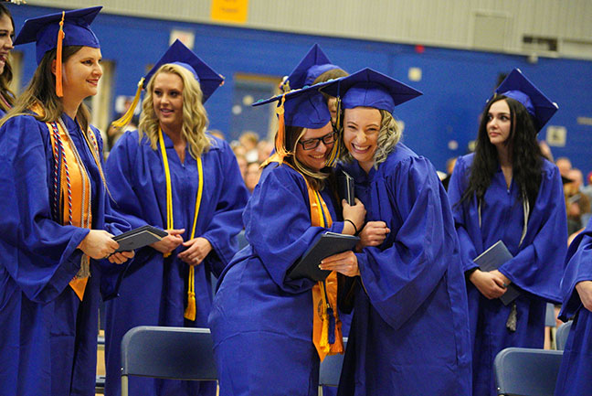 photo of female students at the commencement ceremony hugging in their caps and gowns. They are surrounded by more female student graduates in caps and gowns.