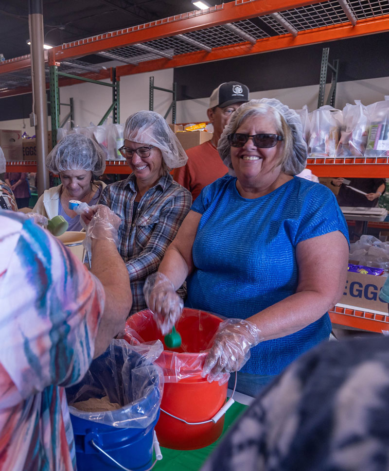 Photo of three volunteers with their hair covered measuring food into mixing containers with shelves in the background