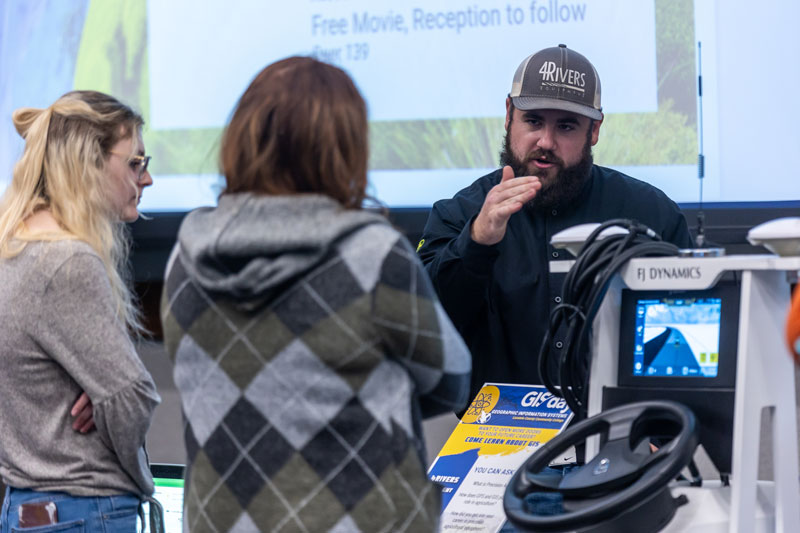 photo of a 4Rivers employee talking to two women at the 2022 GIS Day with a driving simulator in the photo