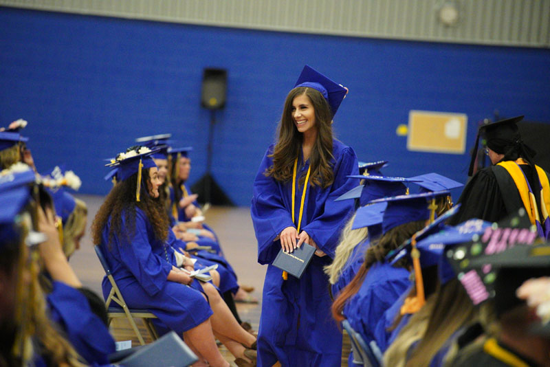 photo of students in caps and gowns at graduation with one student standing holding her diploma