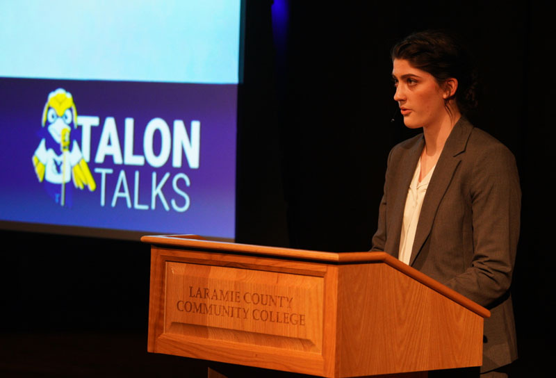 An student on stage speaking for Talons Talks with a screen saying "Talon Talks"