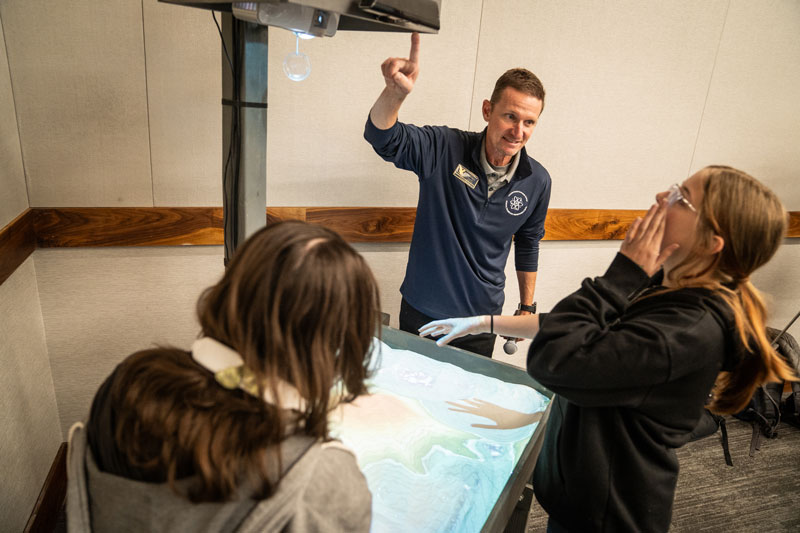 Instructor Trent Morrell talks with two students who are looking at the topographic sand table
