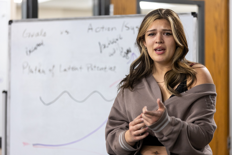 a student speaking in front of a whiteboard with writing on it