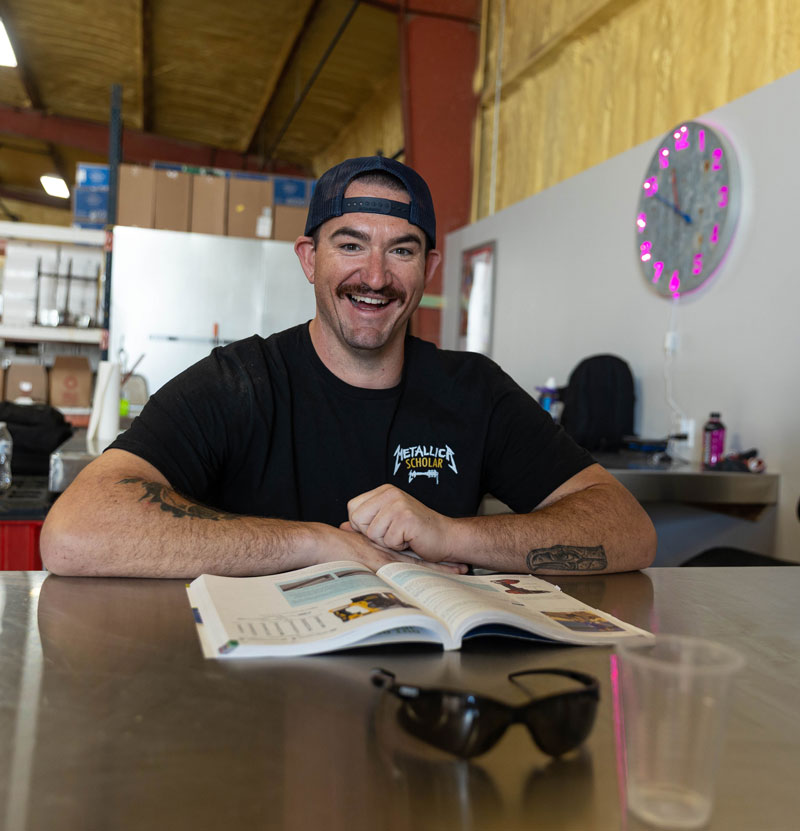 Chase Lilly, a member of LCCC’s first Metallica Scholars Initiative cohort in a backward cap wearing a Metallica Scholar shirt sits at a table with an open book during training at Advanced Comfort Solutions in Cheyenne.