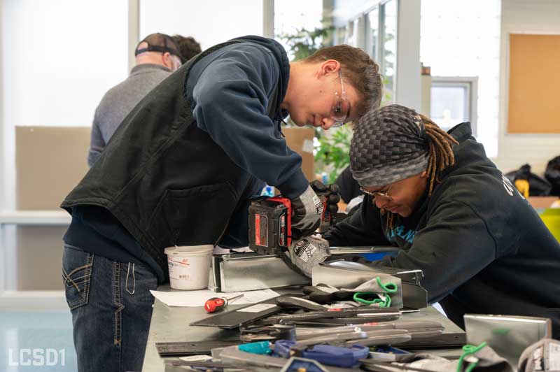 two students are holding and drilling metal pieces on a table with several tools on it.