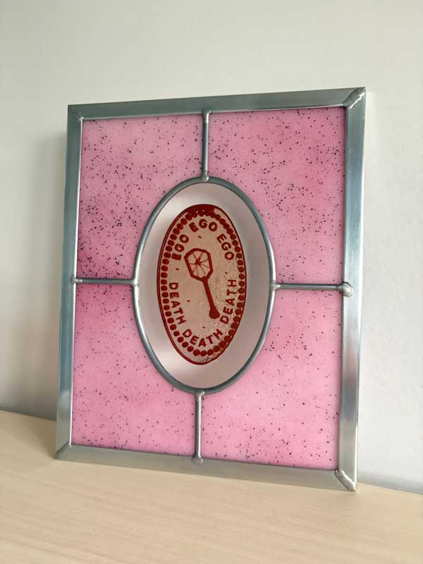 Glass artwork by Jaclyn Miller with four panels joined together with an oval one in the middle. The oval one says "Boo Ego Ego Death Death Death" with a fly swatter drawing in the middle.