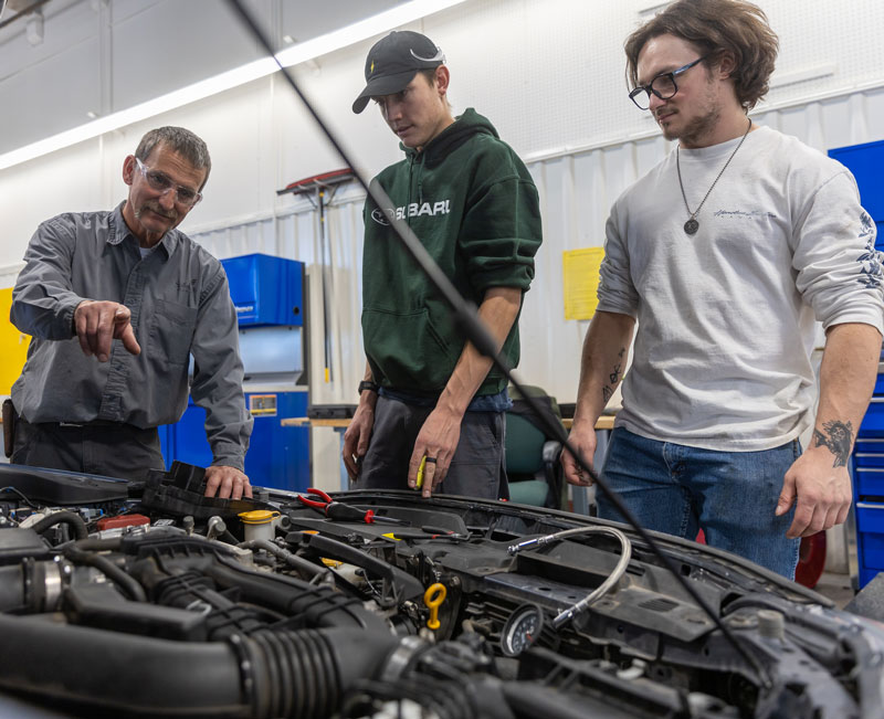 photo of faculty member and two students looking under the hood of a car. Faculty is pointing at something.