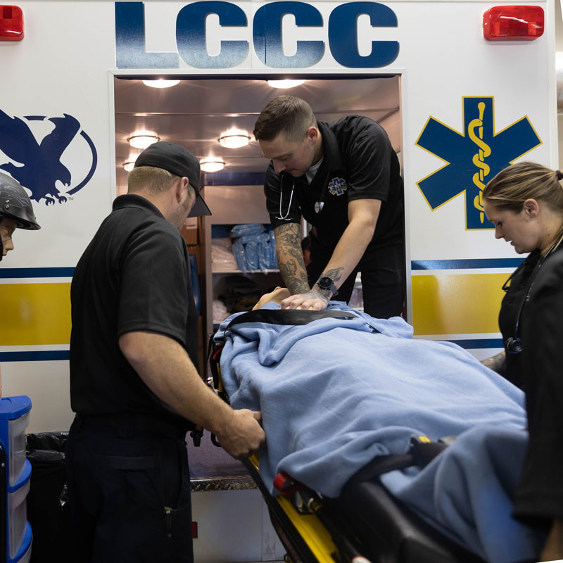 EMS students doing a mock training with an ambulance in the LCCC training area. There is a practice model on the gurney and three students around it.