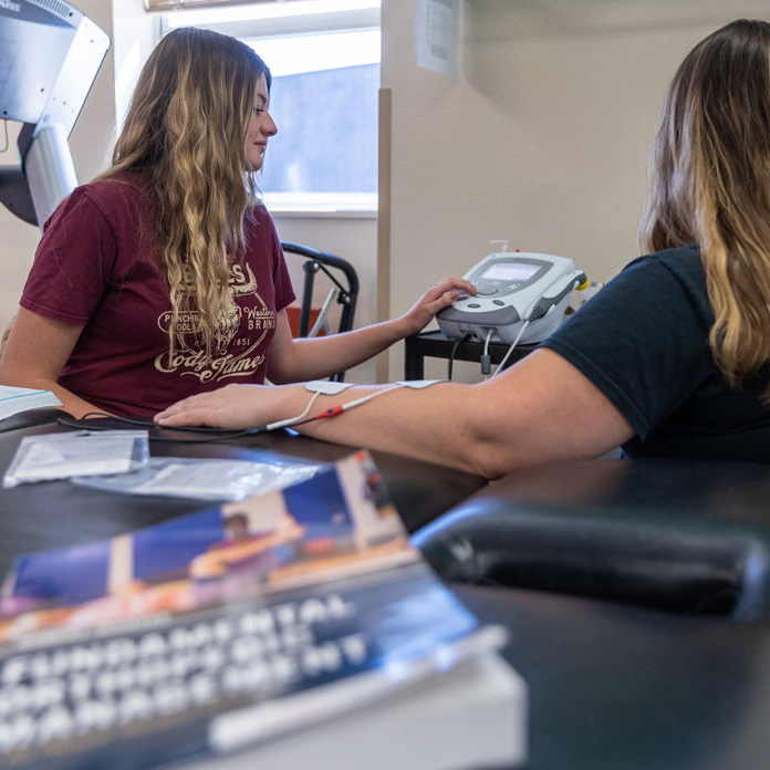 Photo of physical therapy assistant students in class with electric stimulation therapy machine. One student has wires hooked to arm. Other student is looking at the machine.