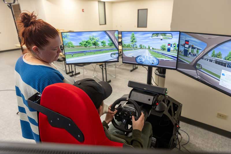 Instructor standing next to a student driver in the driving simulator with his hands on the steering wheel