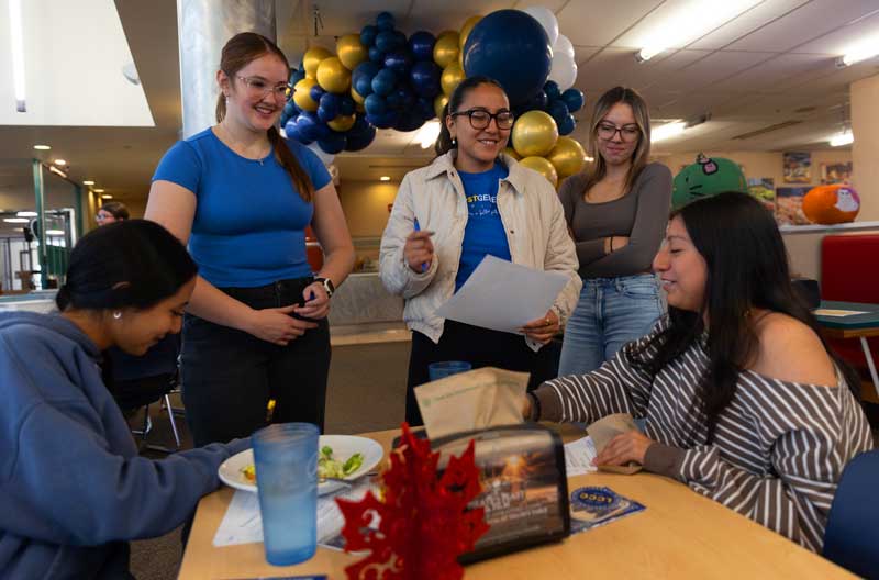 Students in the dining hall for the Davis First Generation lunch celebration. Some students are standing and talking with students seated at a table.