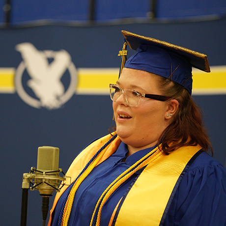 photo of female student at commencement in cap and gown speaking into a microphone on stage